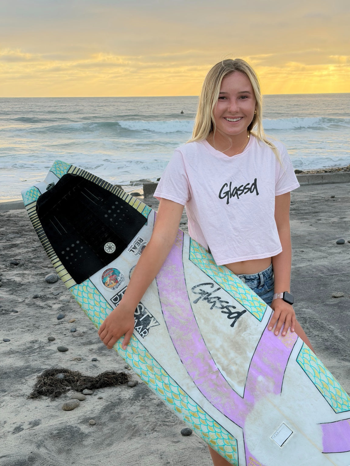 Shiloh Page wearing the pink Glassd Daily Crop Tee with her surfboard at sunset